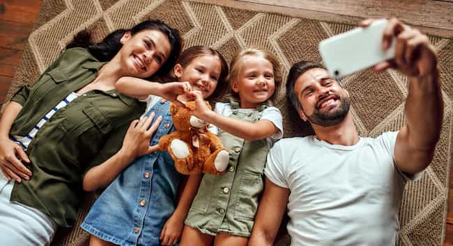 Тато, мама і дві милі доньки лежать на килимі на підлозі у вітальні і роблять селфі на смартфон. Щаслива родина весело провести час разом.