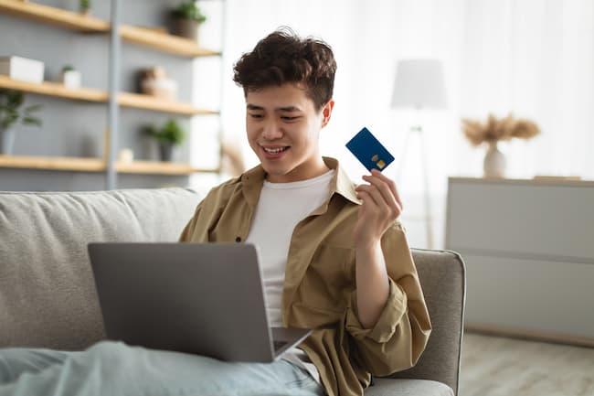 Operación financiera Zdіysnennya. Un retrato de un muchacho asiático sonriente, que muestra una tarjeta de crédito de débito, sentado en el sofá de su casa con una computadora personal en las rodillas, una computadora vicorist y maravillándose con la pantalla.